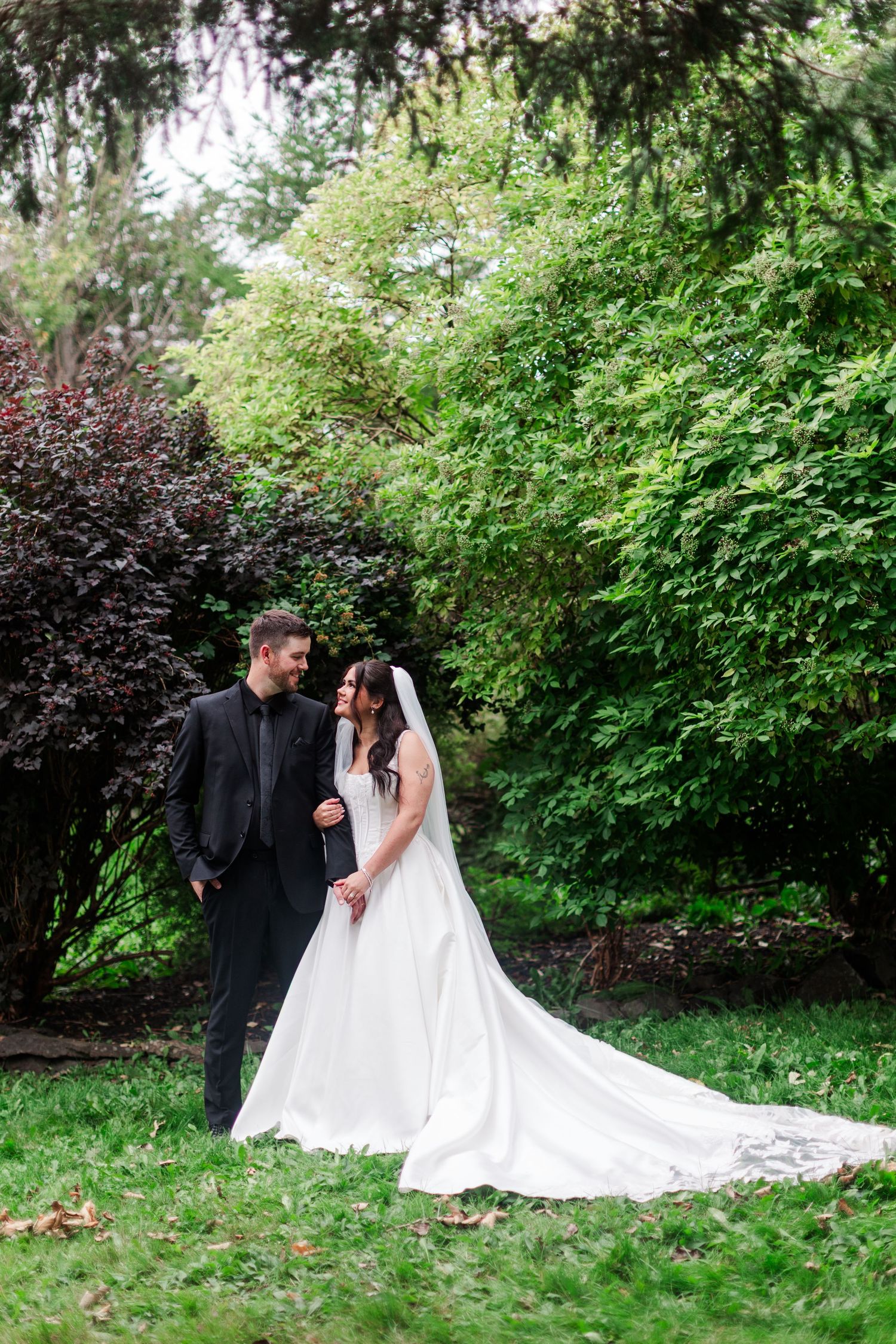 Bride in a white gown with a long train and veil stands beside a groom in a black suit on a grassy lawn surrounded by lush green trees.