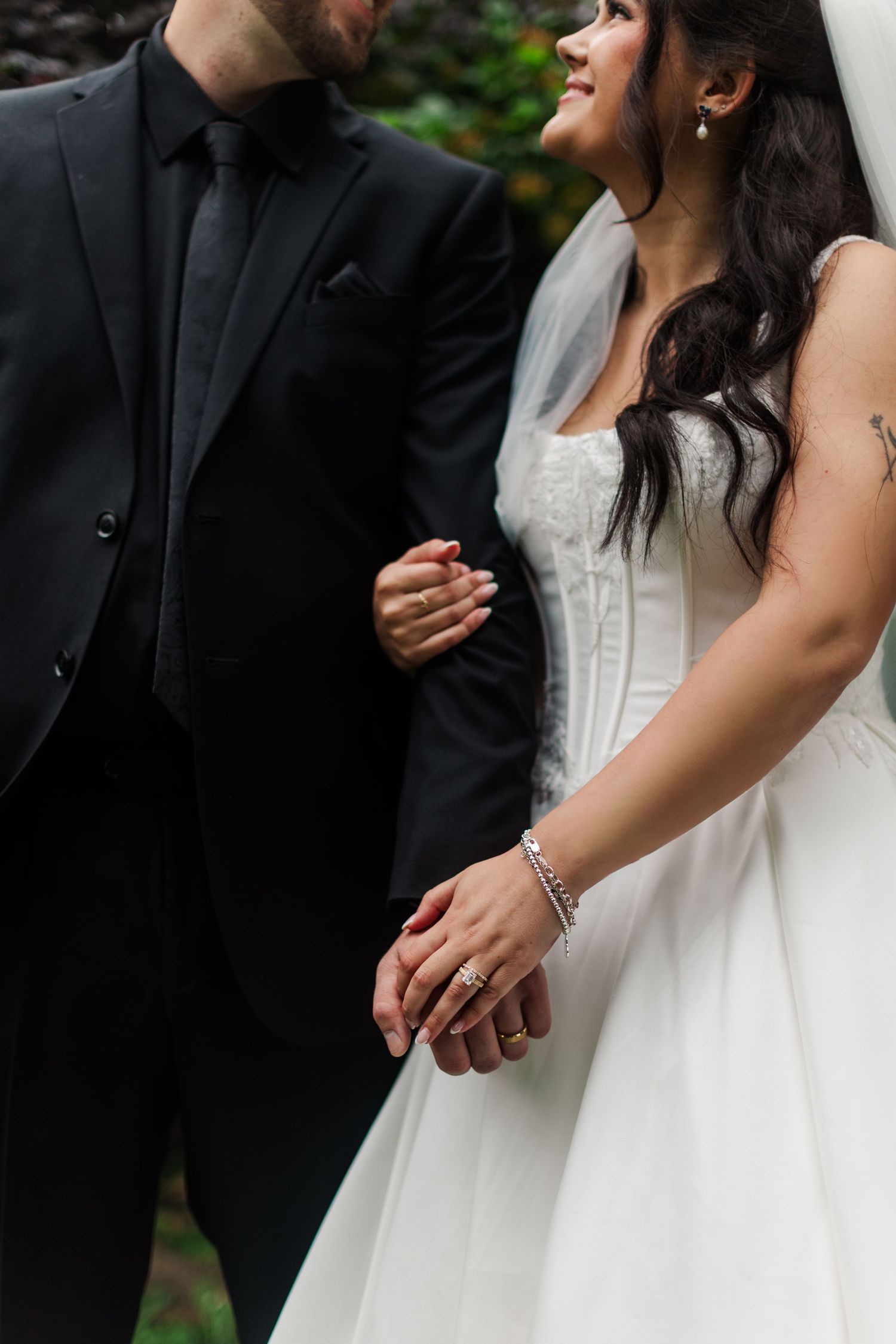 Bride in a white strapless wedding gown and veil holds the arm of a groom in a black suit and tie while smiling outdoors.