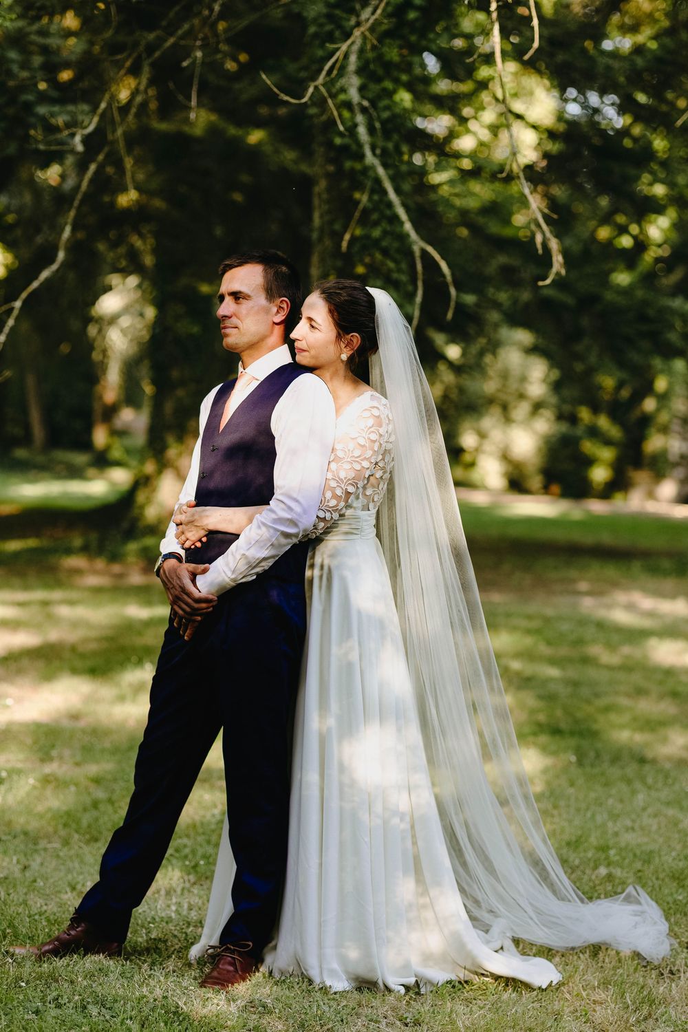 Bride in white veil embracing groom from behind in sunlit outdoor garden setting.