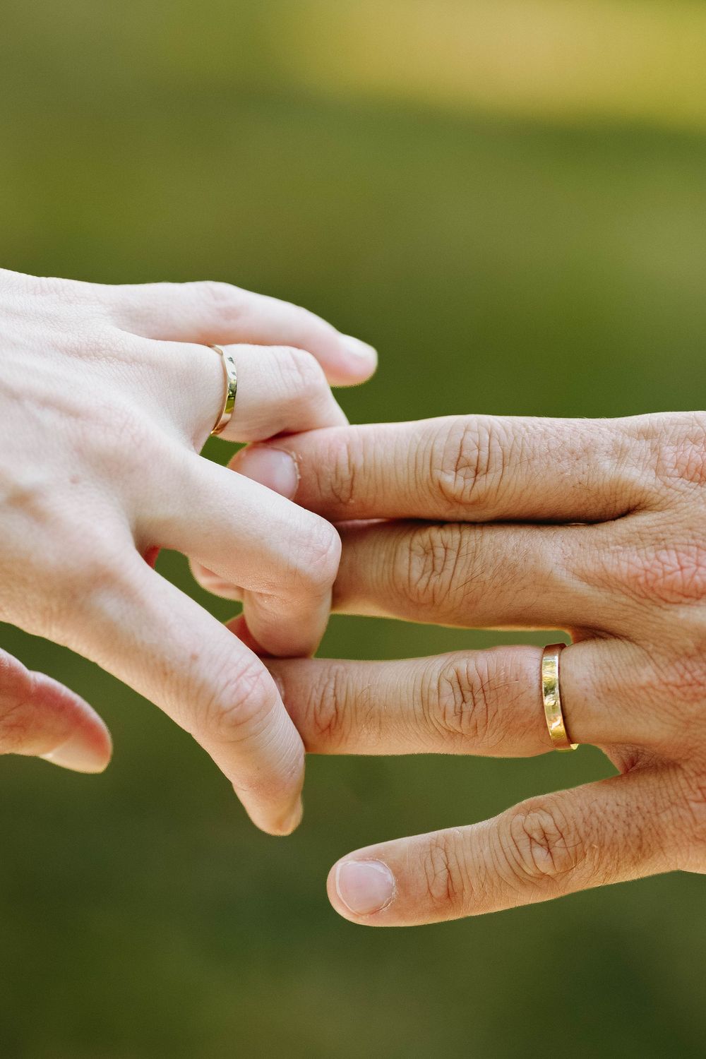 Two hands with matching gold wedding bands intertwined against a green bokeh background.
