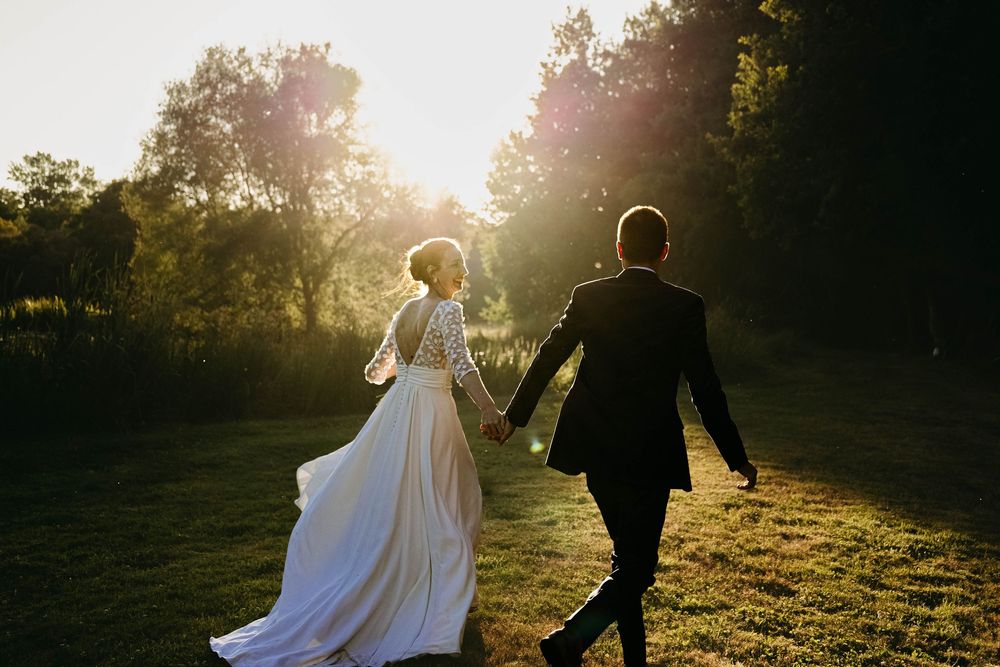 Bride and groom holding hands walking through sunlit garden at golden hour in wedding attire.