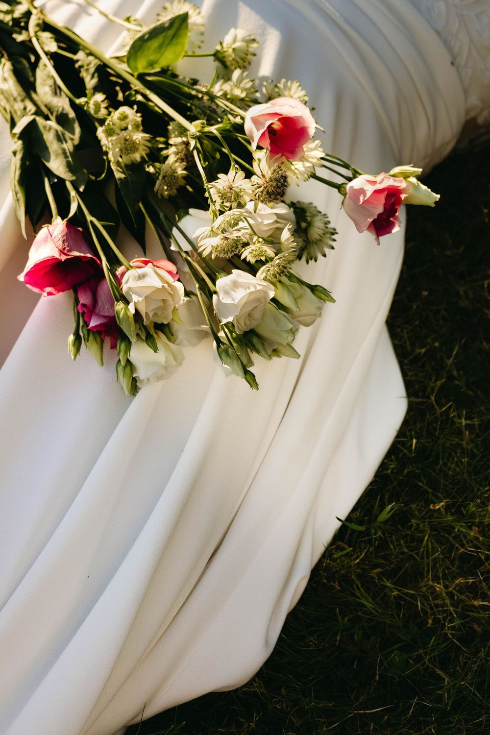 Bridal bouquet of pink and white roses resting on a flowing white wedding dress on green grass.