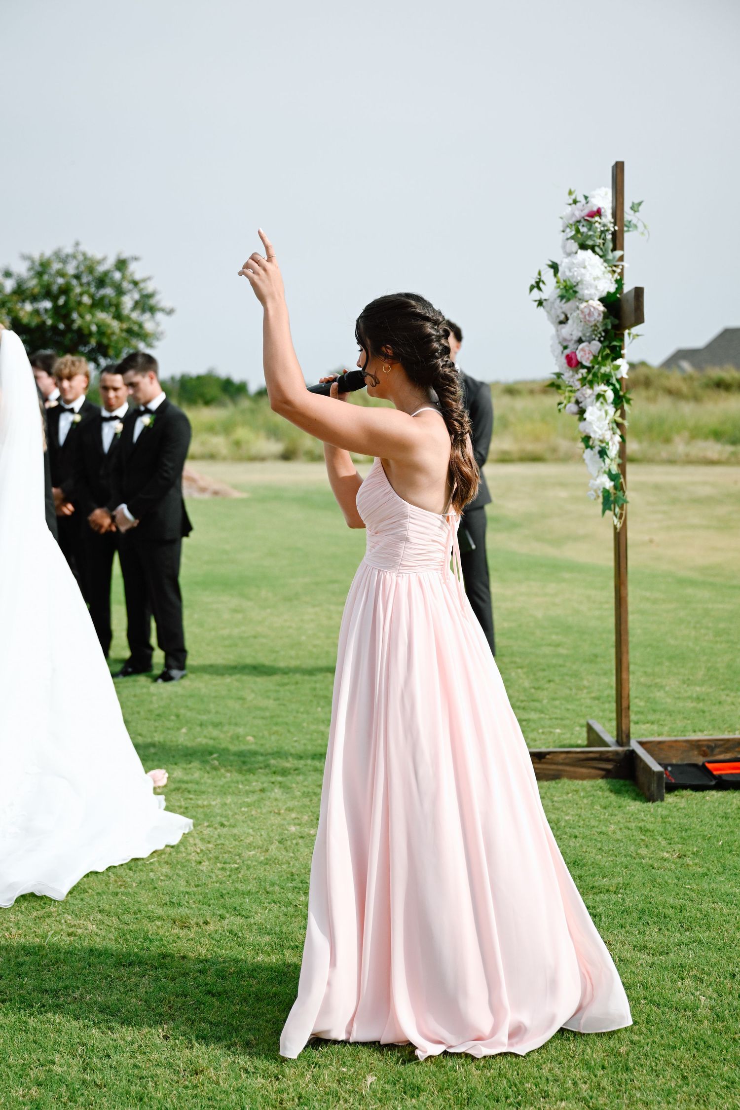 Bridesmaid in pink gown gestures at outdoor wedding ceremony with floral arch and groomsmen on green lawn.
