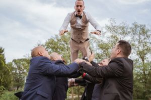Egy világos ruhás személy felugrik, miközben két sötét öltönyös férfi nyúl felfelé alulról egy kültéri környezetben.