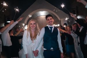 Egy friss házaspár egy boltív alatt áll, miközben a vendégek csillagszórókat tartanak a kezükben egy esti esküvői ünnepségen.