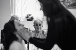 Egy elmosódott sziluett keretezi az előtérben egy idős embert, aki mosolyog a háttérben egy ablakon vagy ajtón keresztül.
