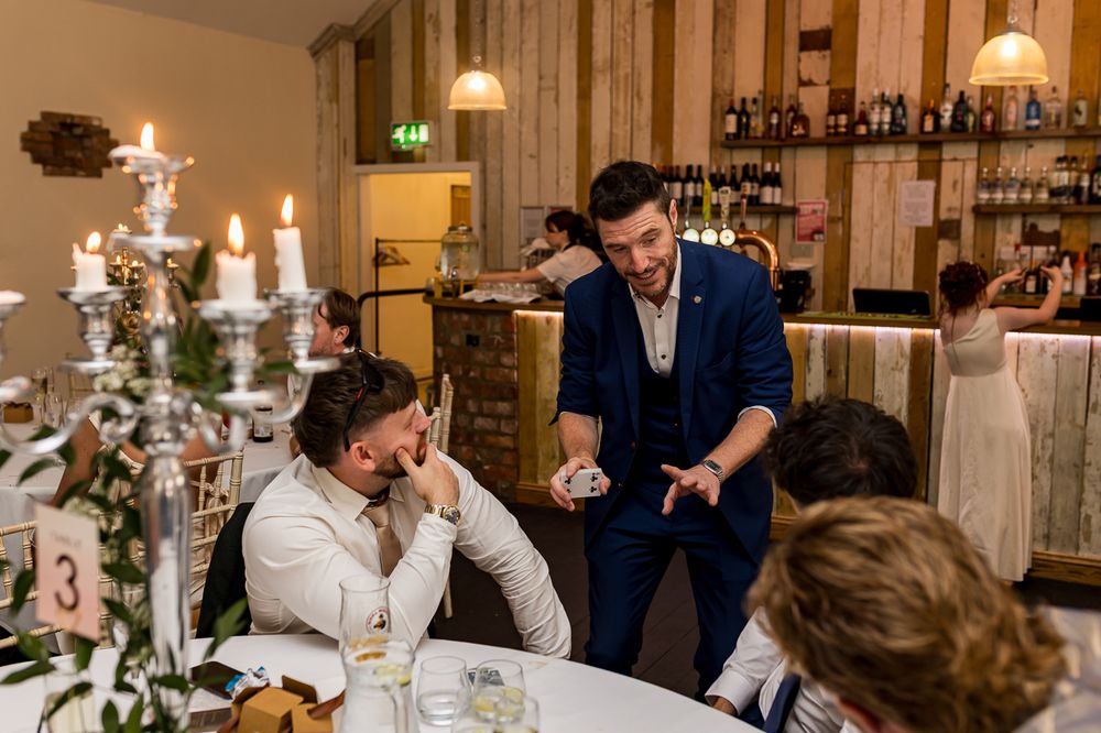A magician performs a card trick for laughing wedding guests at an elegant candlelit reception venue.