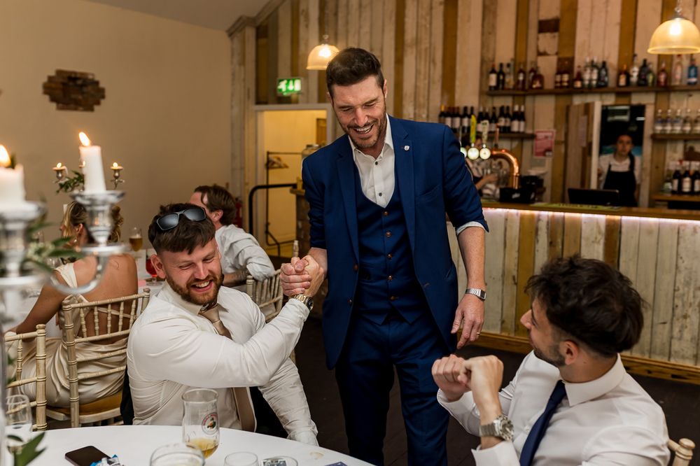 A man in a blue suit shakes hands with guests at a wedding reception dinner table.