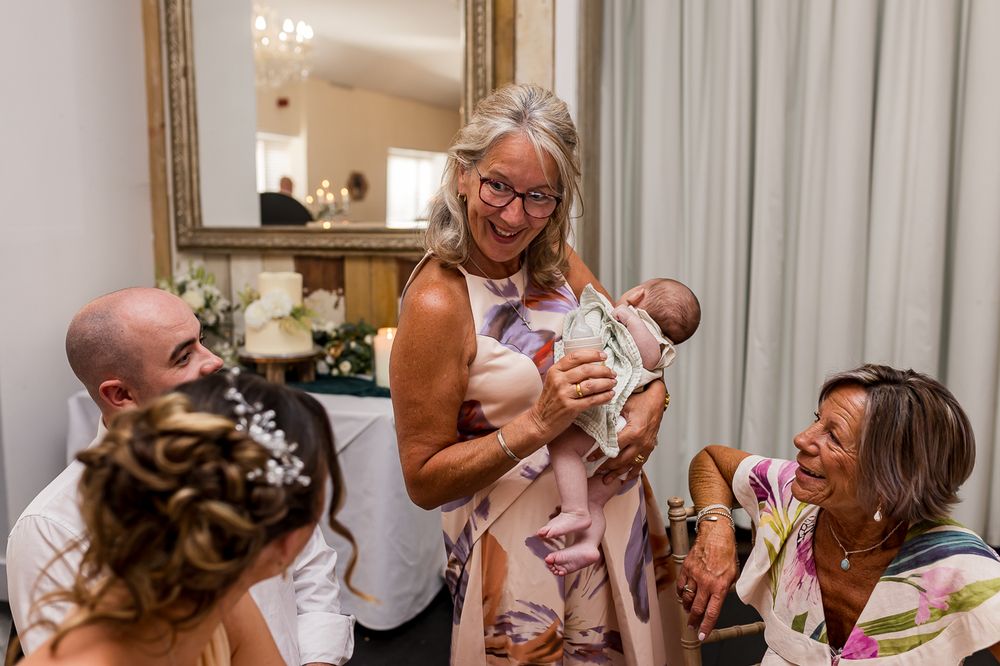 Wedding guests laughing and admiring a newborn baby at a reception, with bride in foreground.
