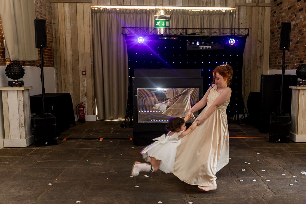 Bride in elegant white gown twirling on dance floor at wedding reception with DJ setup and stage lighting.