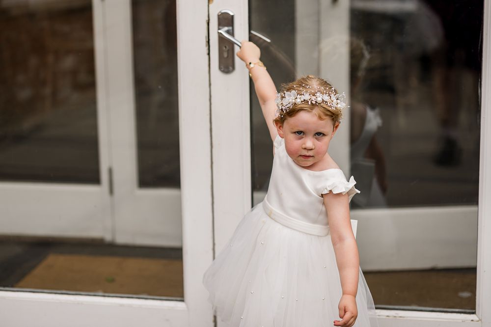 Young flower girl in white tulle dress and floral crown reaching for a door handle at a wedding venue.