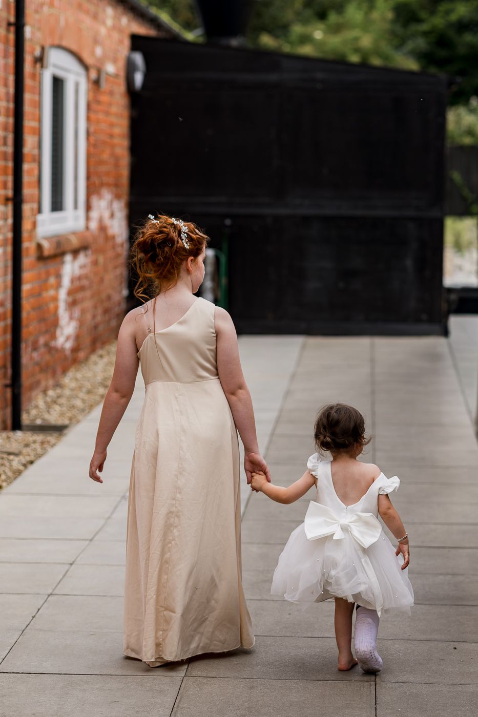 Bridesmaid in champagne gown holds hands with flower girl in white dress walking at outdoor wedding venue.