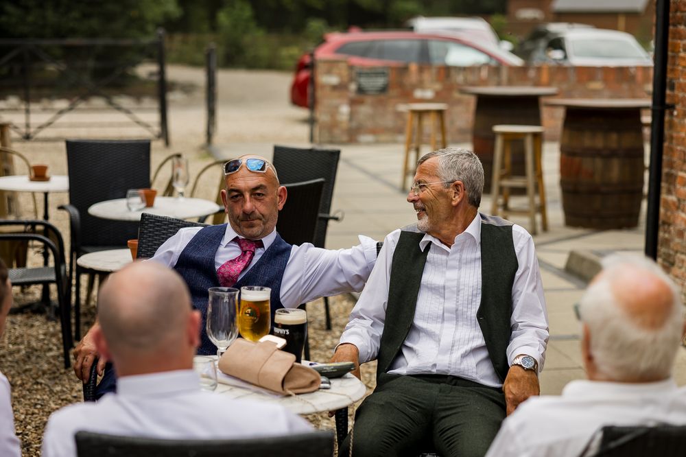Two men in smart attire share a laugh over drinks at an outdoor pub patio table.