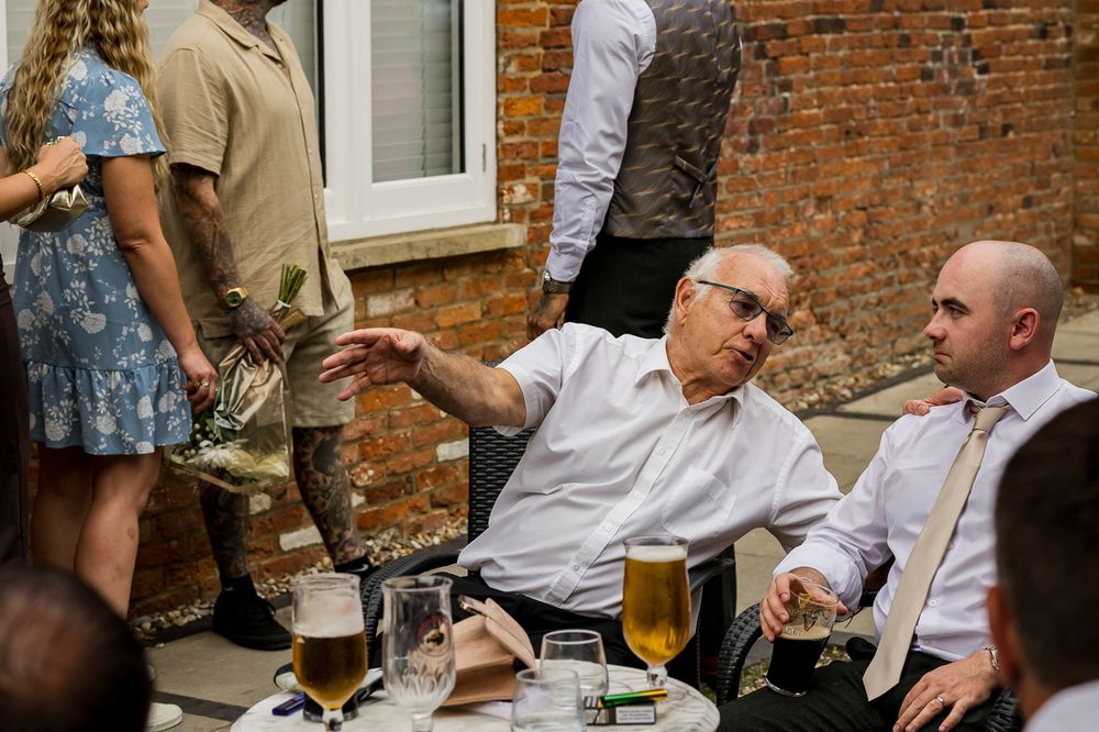 Elderly man reaching across outdoor table with beer glasses at a wedding reception gathering.