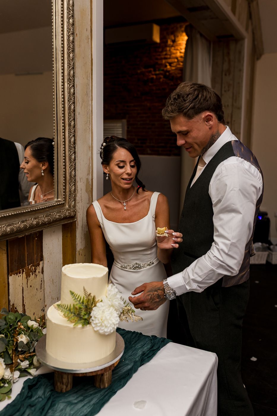 Bride and groom cutting a white two-tier wedding cake decorated with white flowers and greenery at their reception.