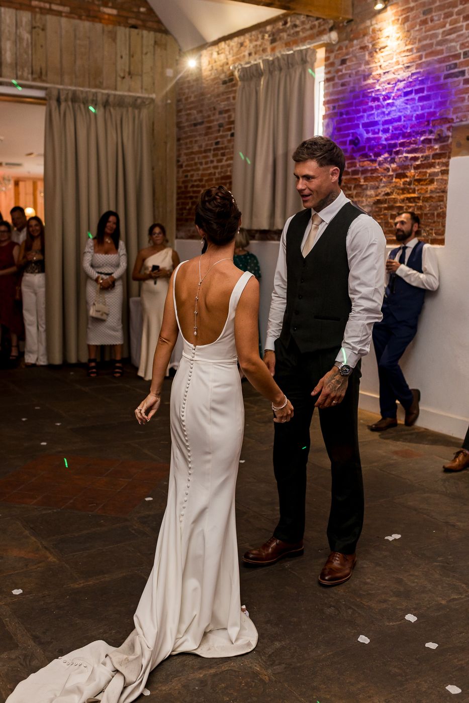 Bride and groom share first dance in rustic brick venue, bride wearing elegant backless white gown with button detail.