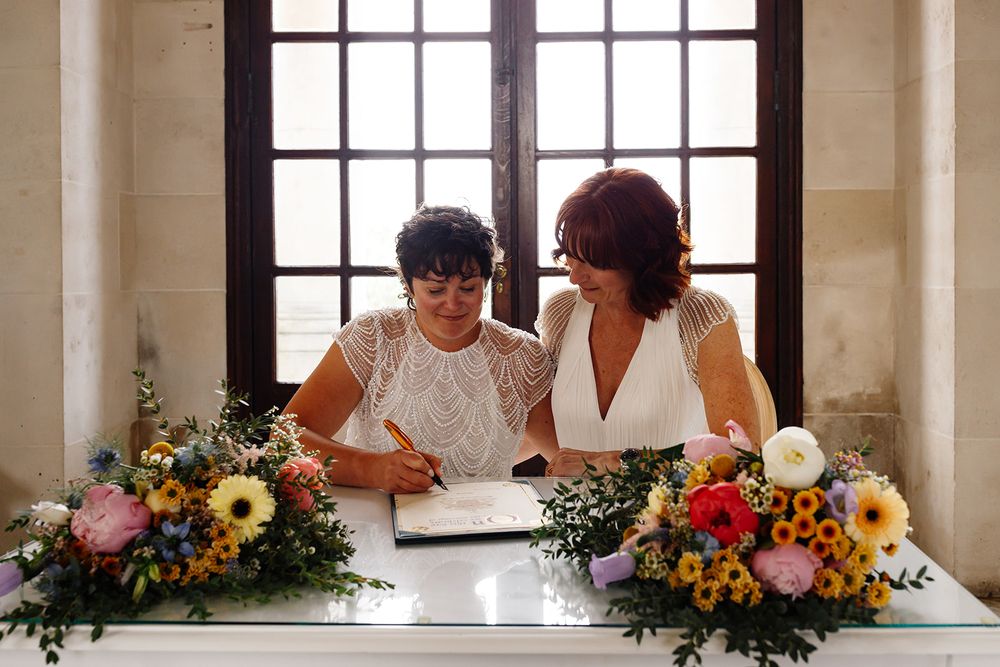 Two brides signing wedding register at decorated table with colorful flowers during civil ceremony.