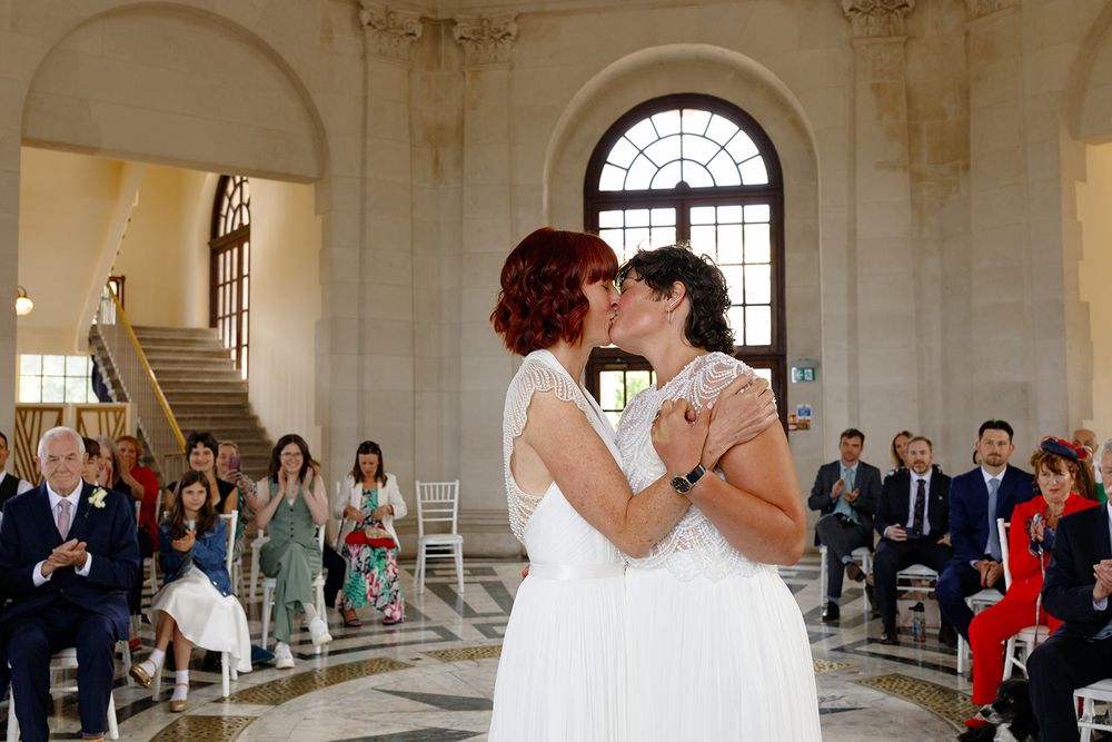 Two brides in white dresses share their first kiss at a same-sex wedding ceremony in an elegant marble hall.