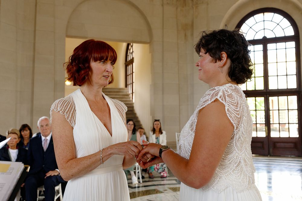 Two brides exchange rings during a wedding ceremony in an elegant venue with guests seated in the background.