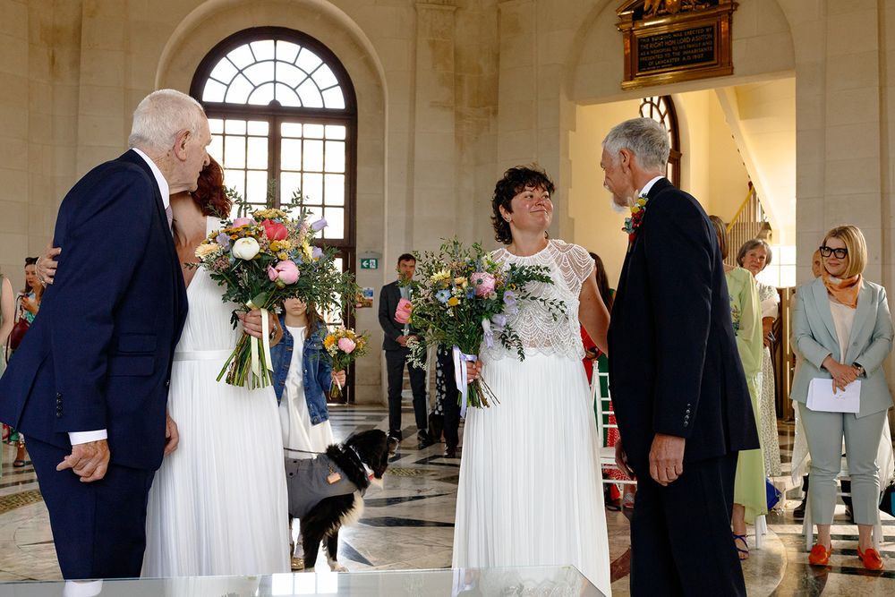 Two brides in white dresses exchange vows with guests and a dog present inside an elegant wedding venue.