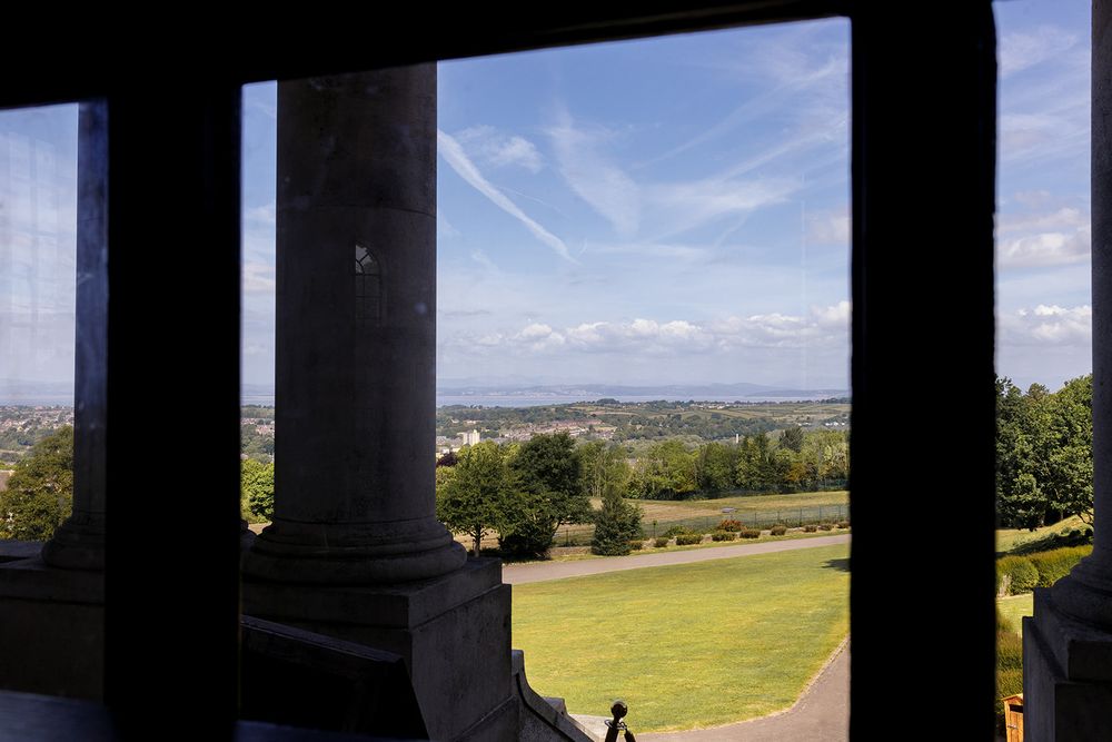View from stone columns of a classical monument overlooking lush green parkland and city skyline under a bright blue sky.