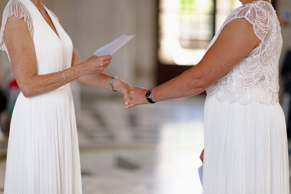 Two women in white dresses exchanging wedding vows, one holding a paper, in a same-sex marriage ceremony.
