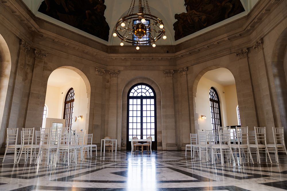 Elegant neoclassical hall with white Chiavari chairs, marble floors, arched windows, and ornate chandelier for events.