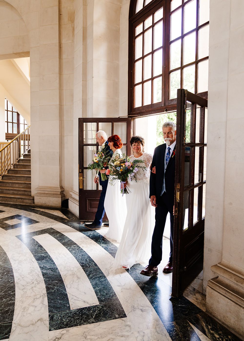 Bride in white gown holding colorful bouquet enters grand marble hall with family at elegant wedding venue.