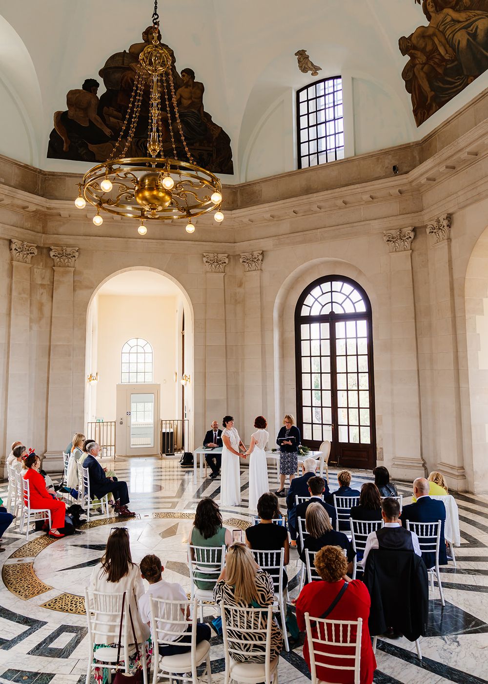 Elegant wedding ceremony in a grand neoclassical rotunda with marble floors, chandelier, and guests seated on white Chiavari chairs.