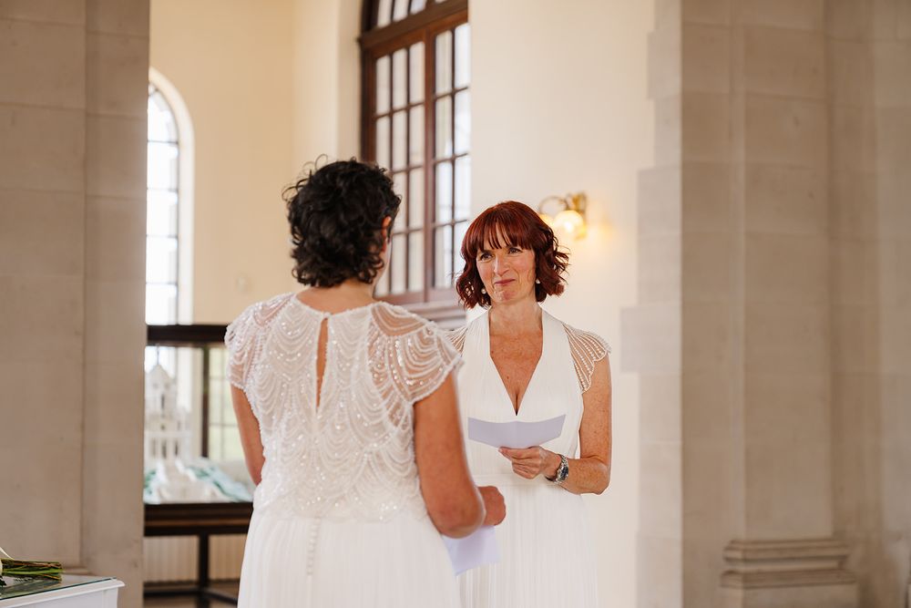 Two women in white dresses exchange wedding vows in an elegant indoor ceremony venue.
