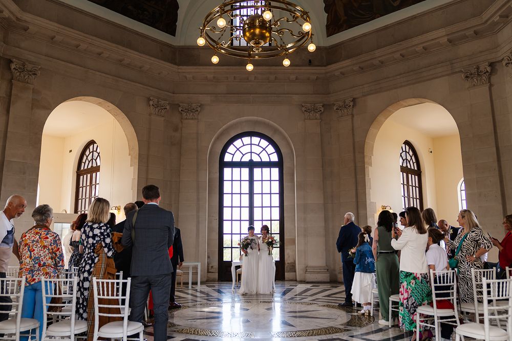 Elegant wedding ceremony in a grand marble hall with arched windows, chandelier, and guests seated in white chairs.