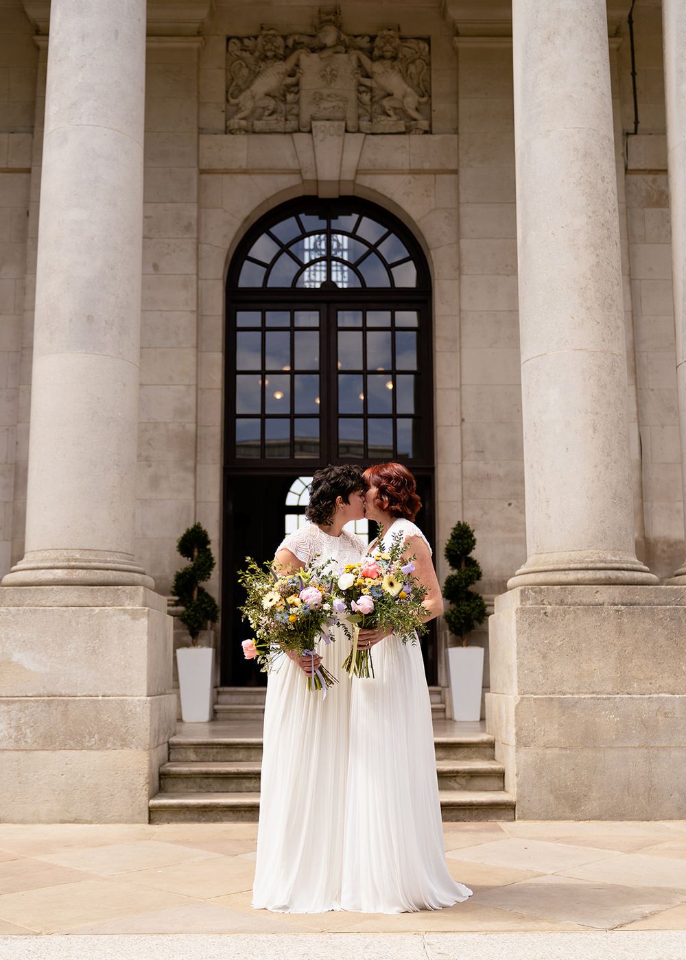 Two brides in white gowns holding colorful bouquets share a kiss outside a grand neoclassical building with stone columns.