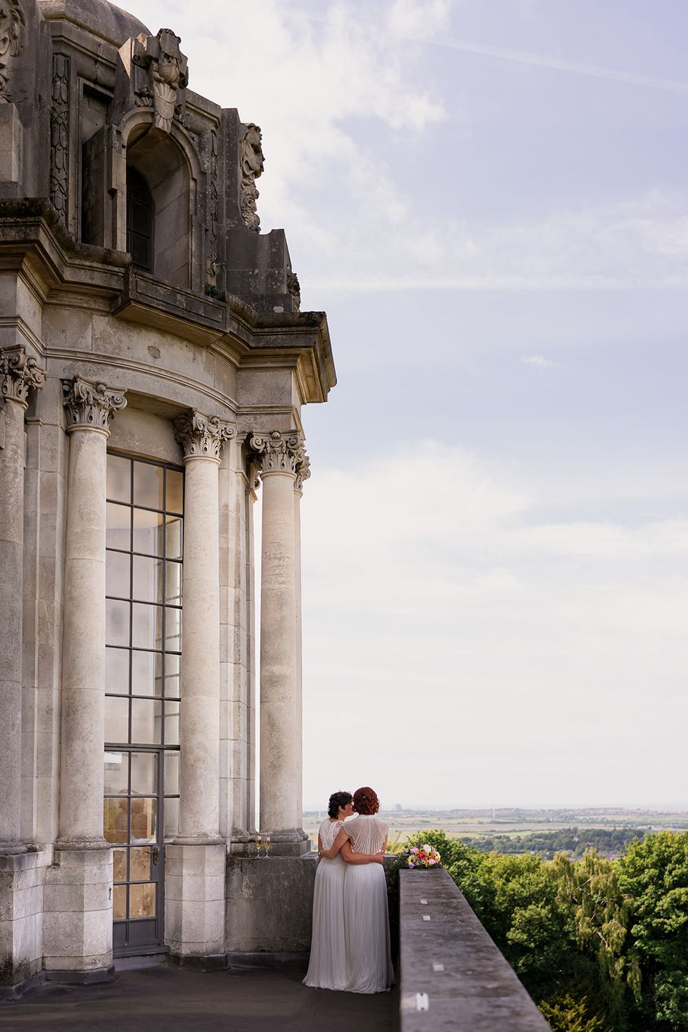 Two brides in white gowns embrace on a grand stone balcony overlooking lush countryside at a château wedding.