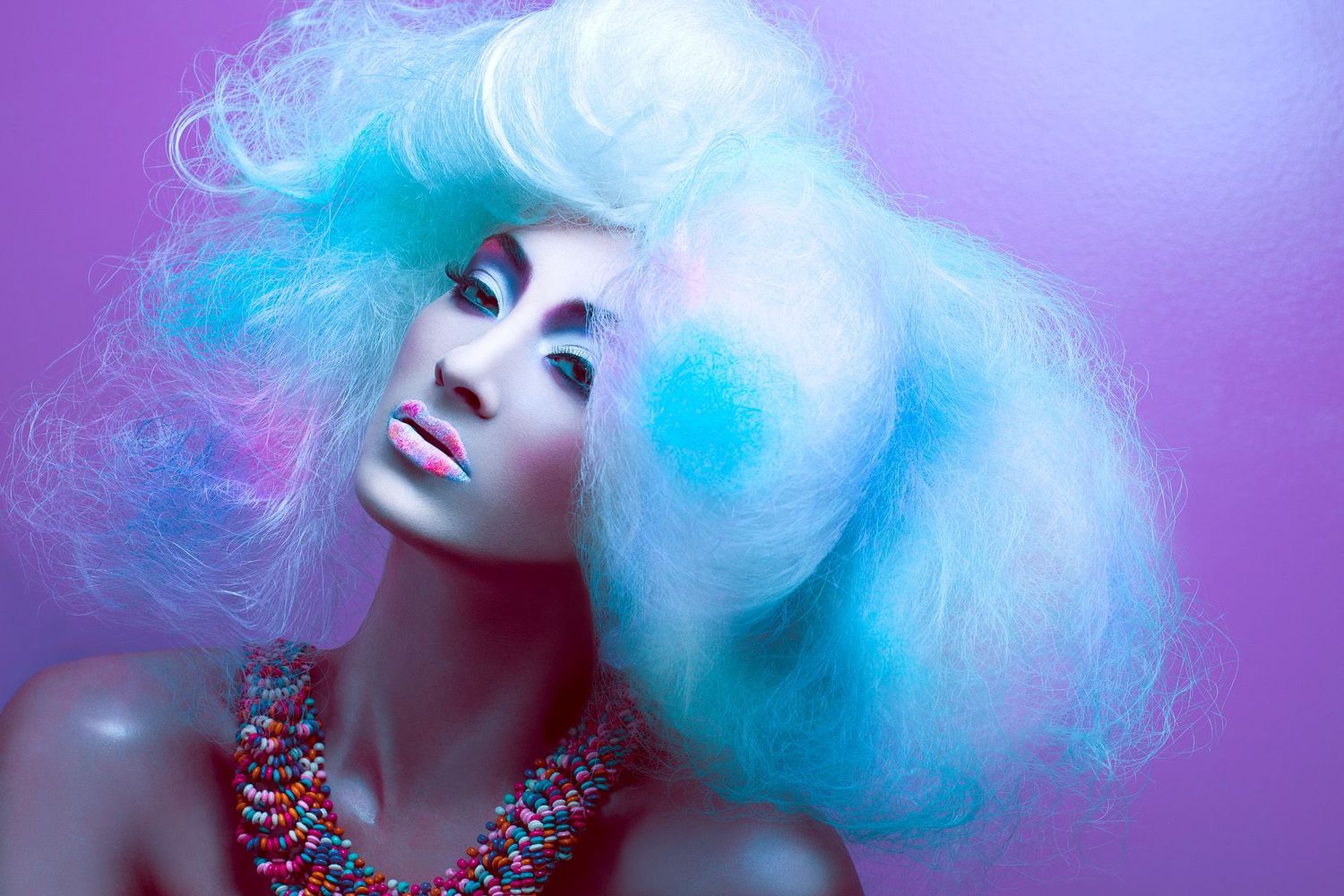 Fashion portrait of a model with voluminous fluffy pastel blue and white hair, a colorful beaded necklace and a vivid purple background.