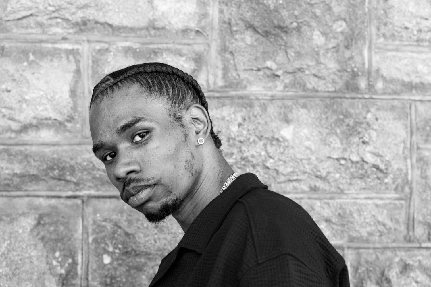 Black and white portrait of a young Black man with braids wearing a black shirt against a stone wall.