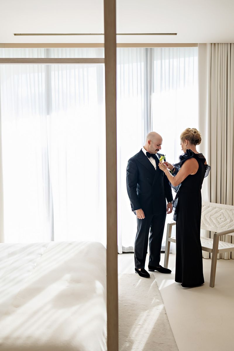 A woman in a black gown pins a boutonniere on a bald man in a tuxedo in a bright, modern hotel room.