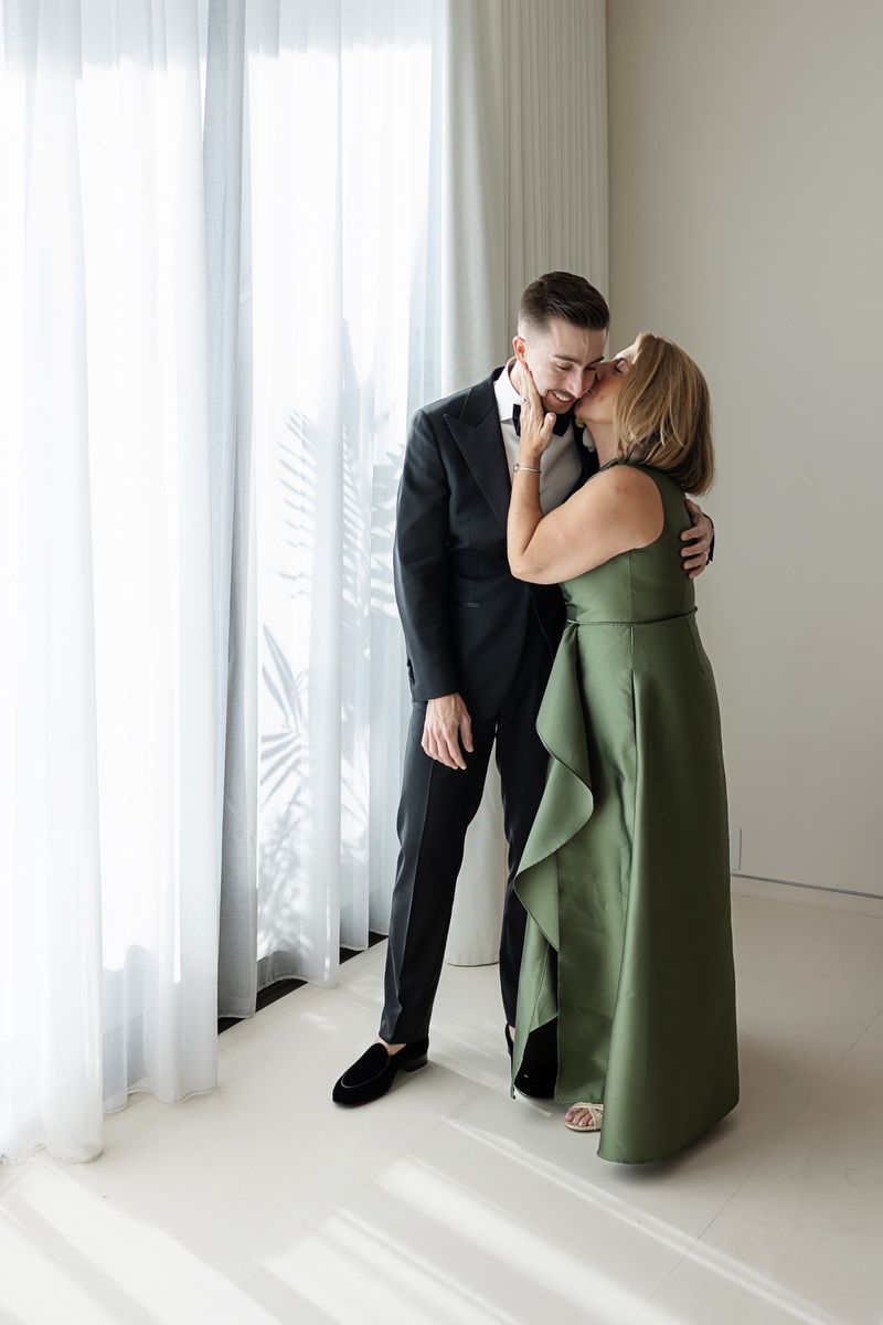 A woman in a green gown kisses a man in a black suit on the cheek near white curtains.