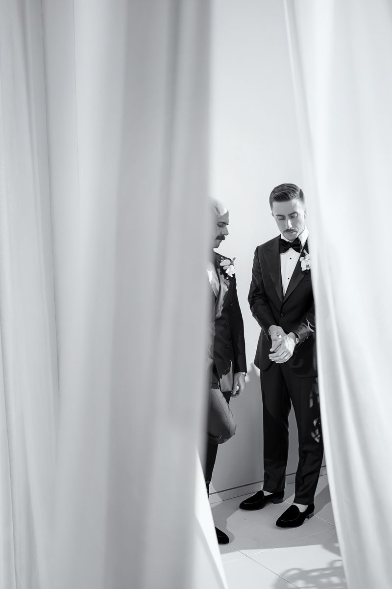 Black and white photo of an elegant couple in formal attire standing between white curtains at a sophisticated event.