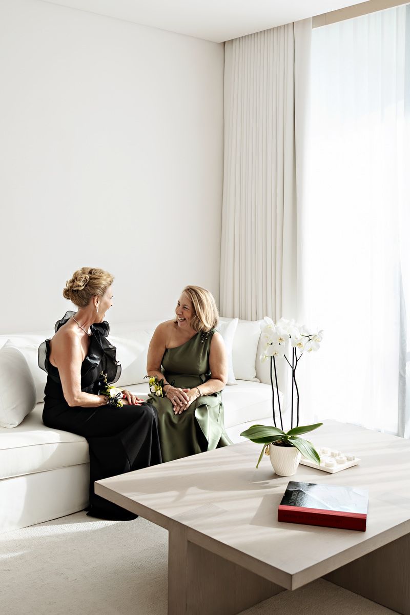 Two women in elegant dresses sit conversing on a white sofa beside a marble table with orchids.