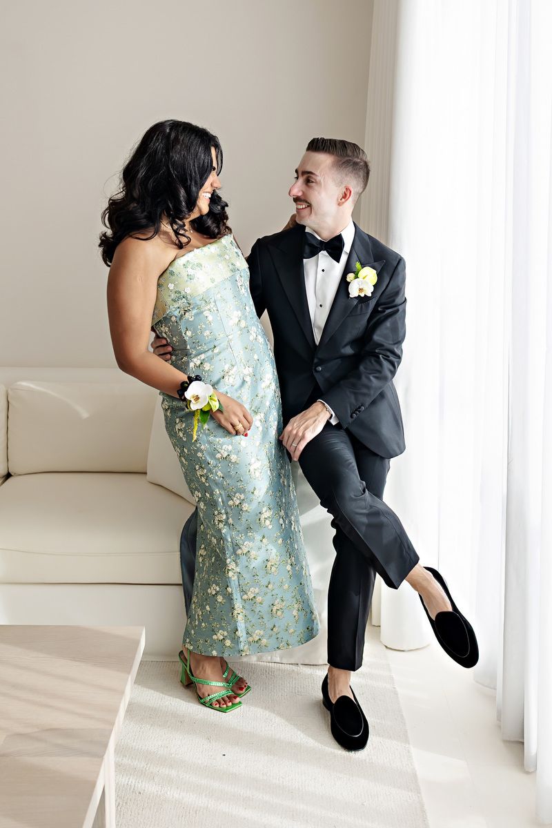 Couple in formal attire, woman in floral gown and man in tuxedo, posing together in elegant white room.
