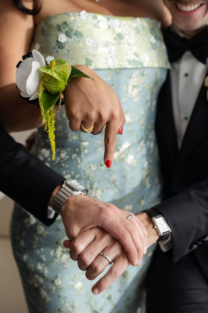 Couple in formal attire holding hands, woman wearing floral wrist corsage and red nails, man in tuxedo with silver watch.