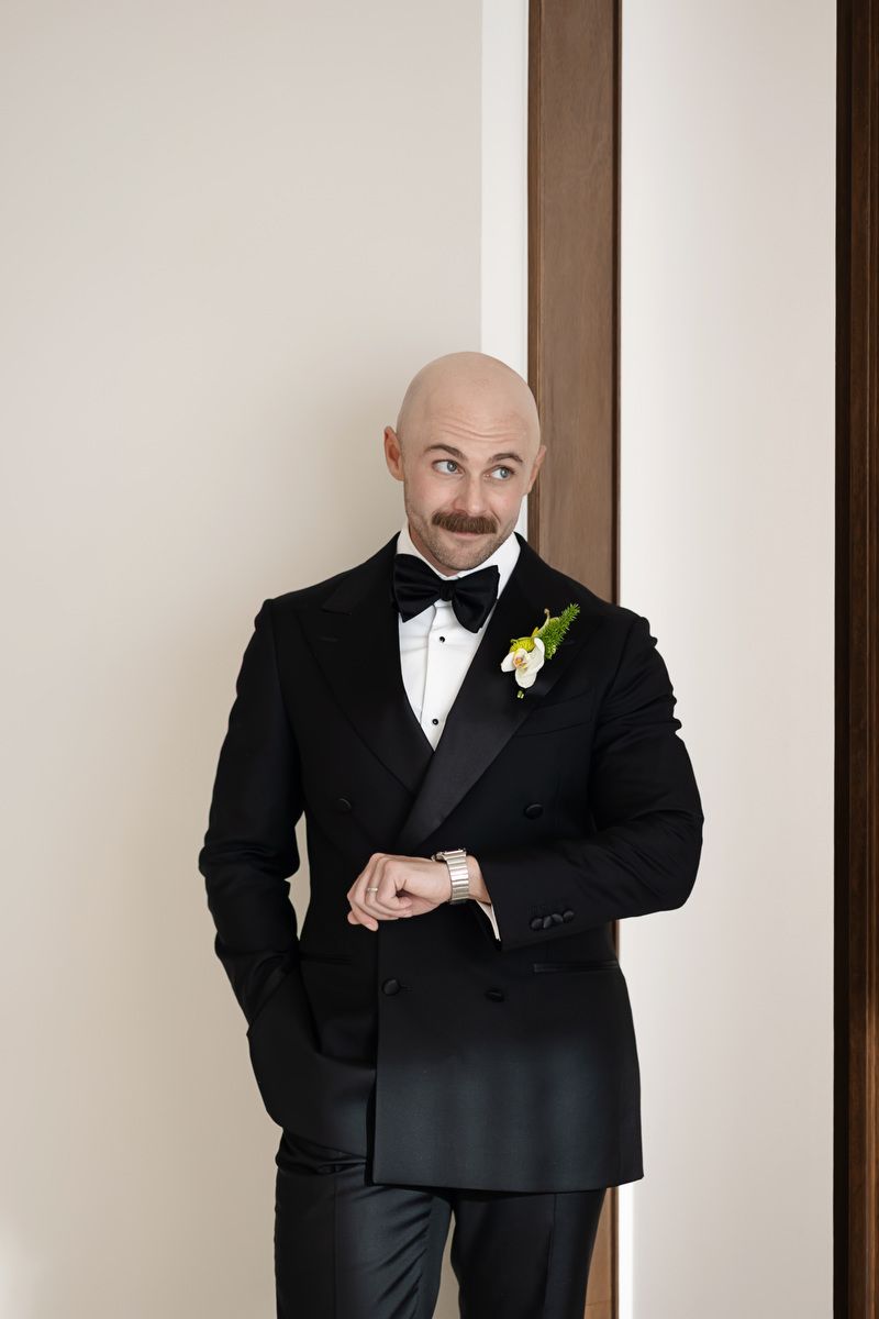 Bald groom in black tuxedo with bow tie and boutonniere checks his watch while leaning against a wall.