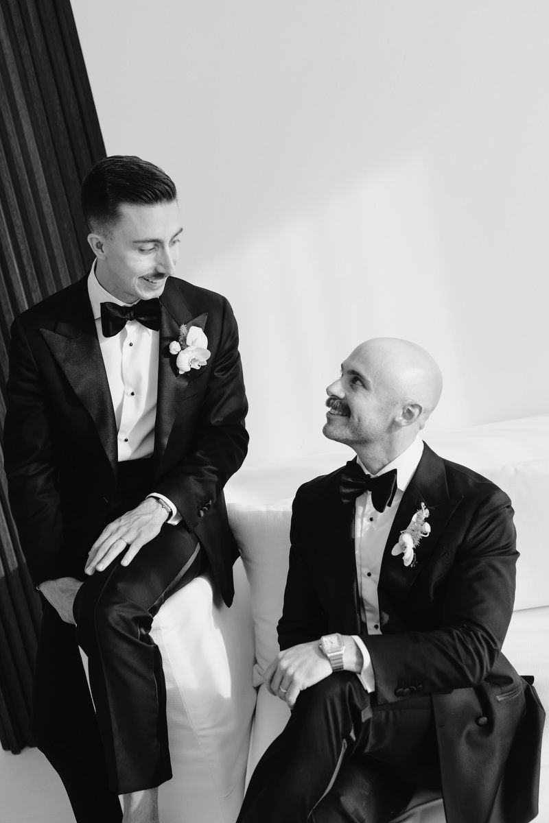 Two grooms in tuxedos with boutonnieres smile at each other while seated together in a black and white wedding photo.