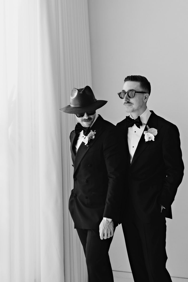 Two grooms in black tuxedos with boutonnieres, one wearing a hat and face paint, pose in a black and white wedding photo.