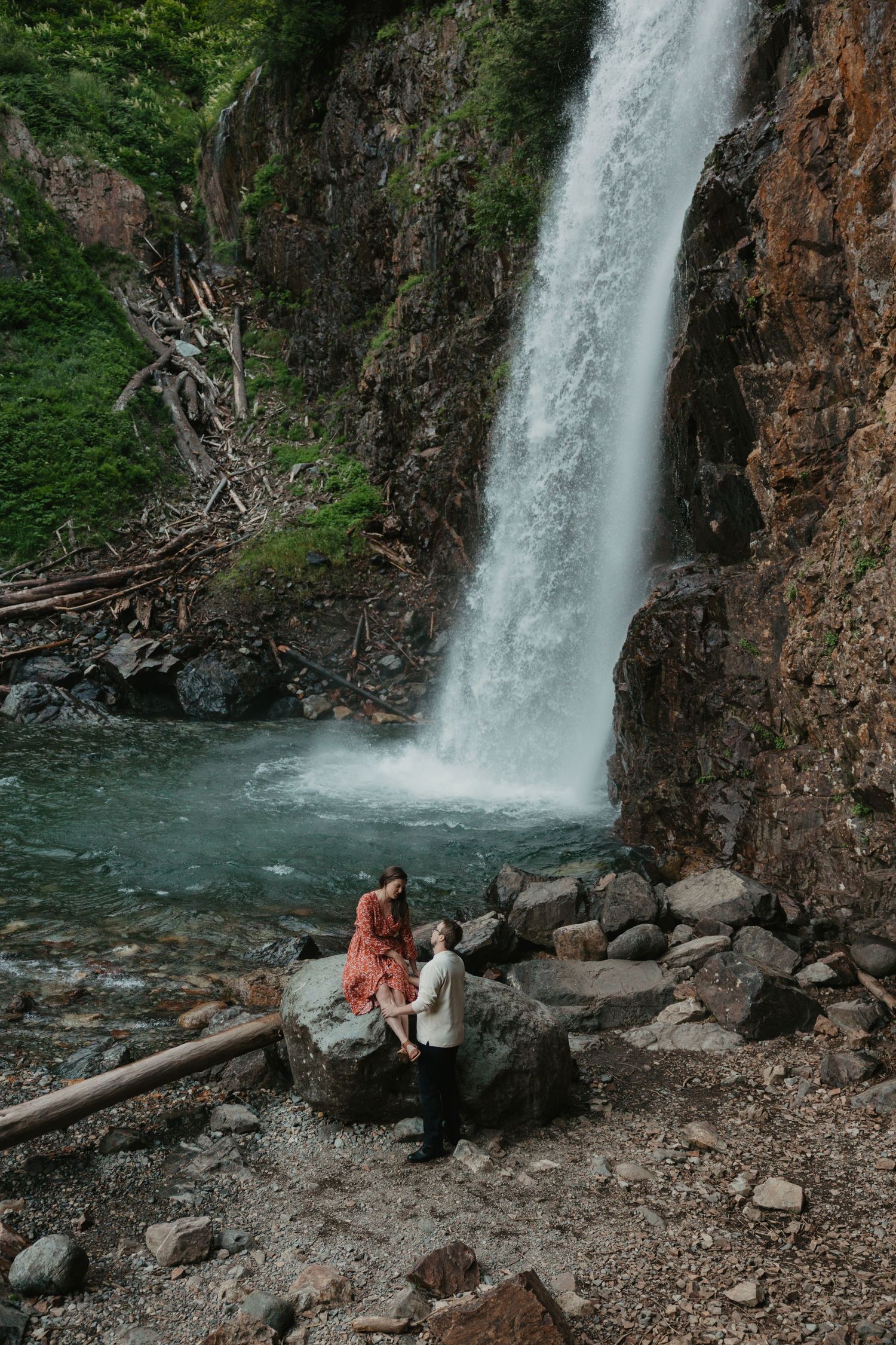 Couple holding hands on rocks before a stunning waterfall surrounded by lush greenery and rugged cliffs in Mt. Baker-Snoqualmie forest
