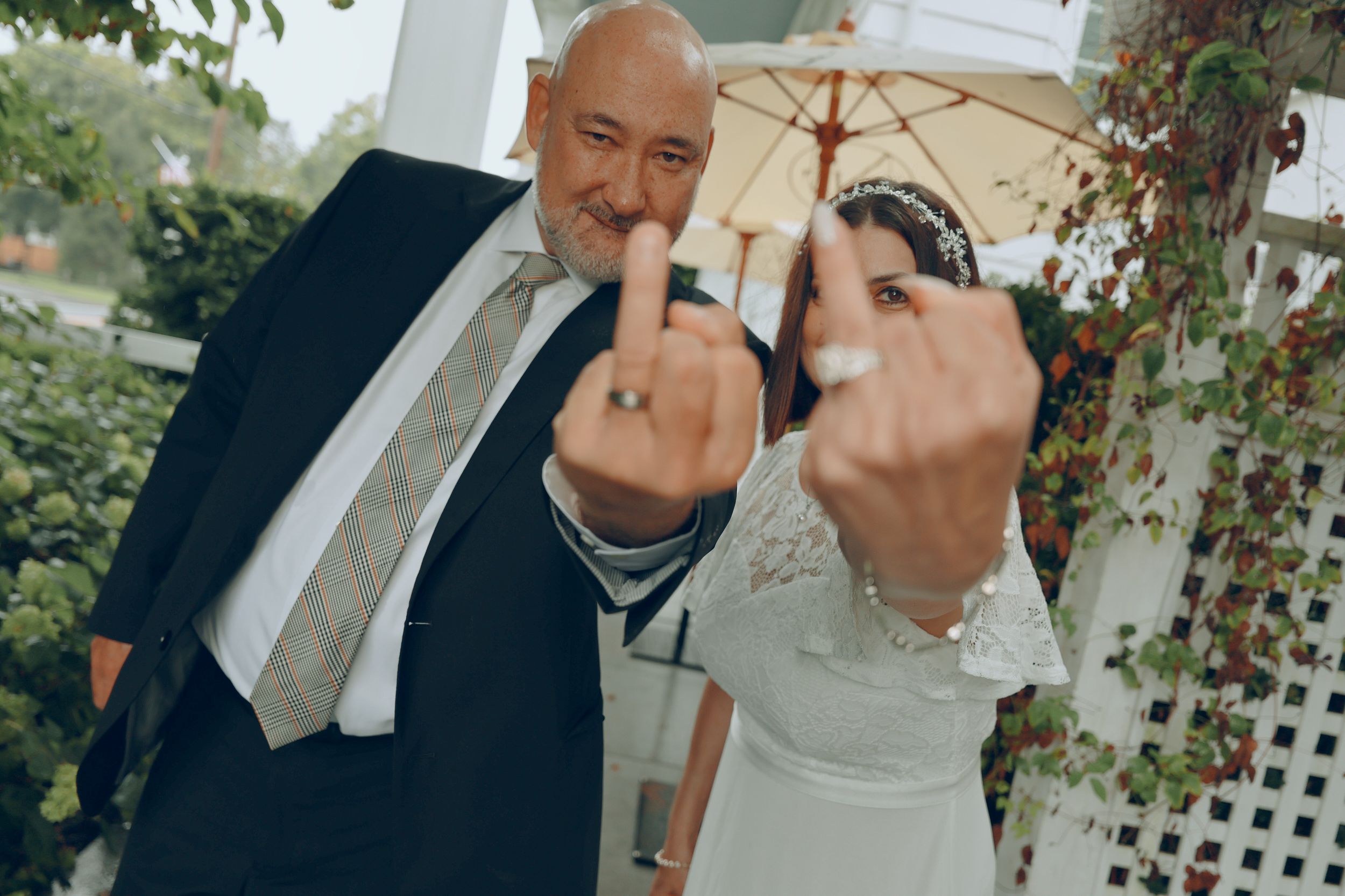 A bald groom in a dark suit and a bride in a lace dress playfully show their rings while raising middle fingers at an outdoor wedding.