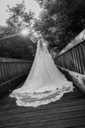 Black and white photo of a bride in a lace-trimmed gown with a long cathedral veil standing on a wooden bridge at sunset.