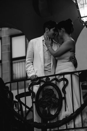 Black and white photo of a romantic couple embracing on an ornate wrought iron balcony, the man in a light suit and the woman in a gown.
