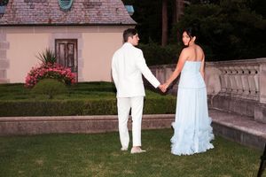 Couple holding hands in white and pale blue formal attire on a manicured garden lawn near a stone balustrade and pink flowers.