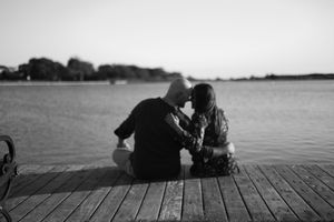 Black and white photo of a couple sitting close together on a wooden pier, embracing as they gaze out over calm water at sunset.